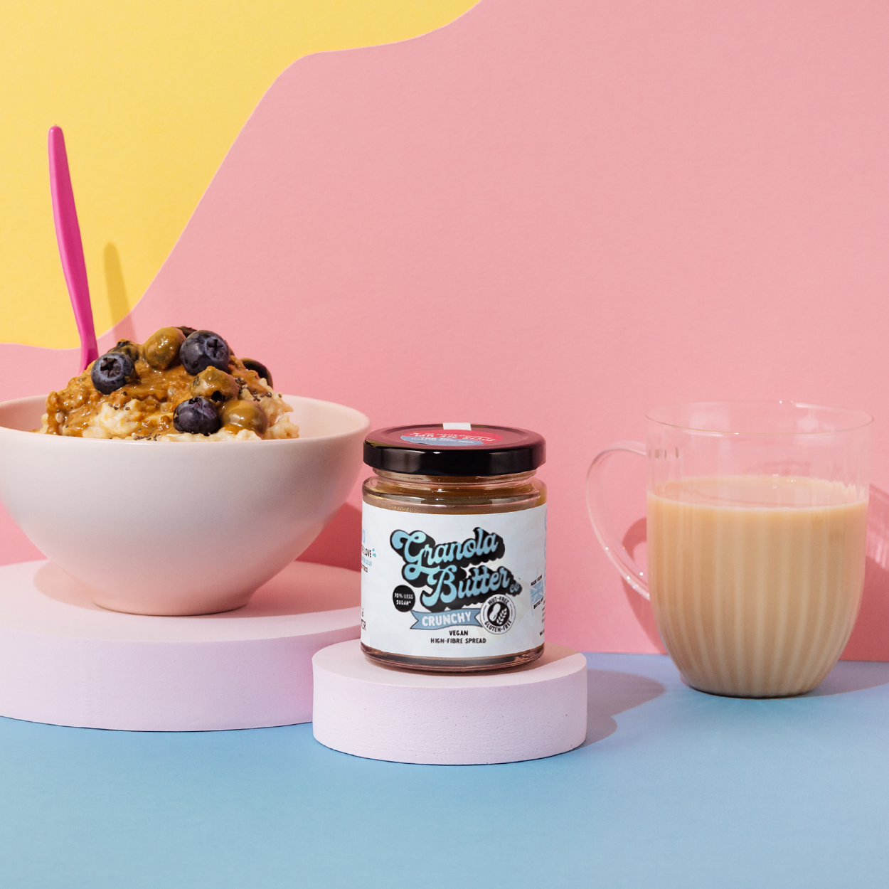 Breakfast bowl or porridge bowl topped with crunchy granola butter and blueberries. A jar of Crunchy Granola Butter and a transparent cup of coffee are also in the image. 